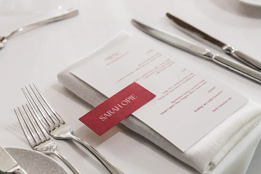 Burgundy wedding place card printed in white, laid across a menu at a set table